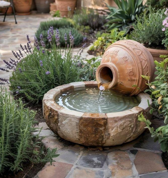 Inspiration image: compact Mediterranean garden with terracotta urn pouring into a stone basin, irregular stone paving, lavender, rosemary and trailing groundcover with pink blooms
